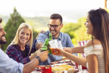 Bir grup genç arkadaş dışarıda barbekü yapıyor, masada otururken kadeh kaldırıyor, bira içiyor ve eğleniyorlar.