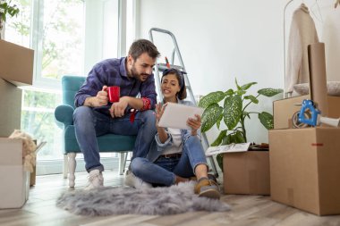 Birbirine aşık bir çift, tablet bilgisayar ve dergiler kullanarak ev dekorasyonu fikirleri ararken kahve içip eğleniyor.