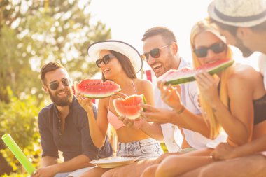 Havuz başında bir yaz partisinde bir grup arkadaş, yüzme havuzunun kenarında oturmuş, soğuk karpuz dilimleri yiyor ve eğleniyorlar.