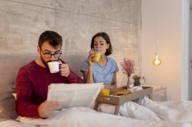 Güzel, genç aşık bir çift pijama giyiyor, sabahları yatakta oturuyor, gazete okuyor, kahve içiyor ve kahvaltı yapıyor, evde dinleniyor.