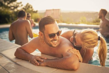 Havuz başında bir yaz partisinde aşık bir çift yüzme havuzunun kenarına yaslanıyor, güneşleniyor ve eğleniyor.