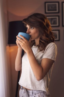Pijama giyen, oturma odasının penceresinin yanında duran, kahve içen güzel genç bir kadın.
