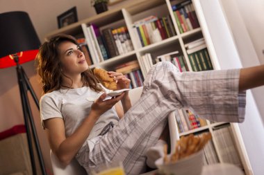 Güzel genç bir kadın pencerenin yanındaki sandalyeye oturmuş kahvaltıda taze kruvasan ve bir bardak portakal suyu içiyor.