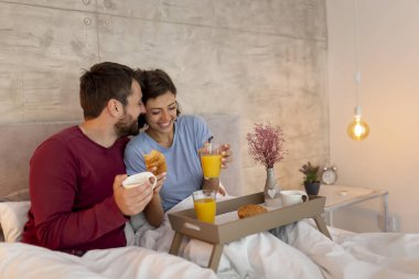 Güzel, genç aşık bir çift pijama giyip yatakta oturuyor, kahve içiyor, kahvaltı yapıyor ve sabah evde dinleniyor.