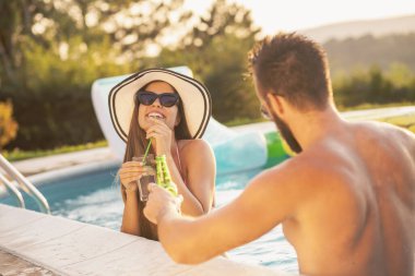 Havuz başında bir yaz partisinde aşık bir çift yüzme havuzunun kenarında durmuş bira içiyor ve eğleniyor.