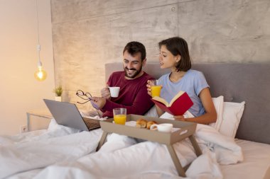 Güzel, genç ve aşık bir çift yatakta kahvaltı ederken internette kitap okuyup sörf yaparken eğleniyor.