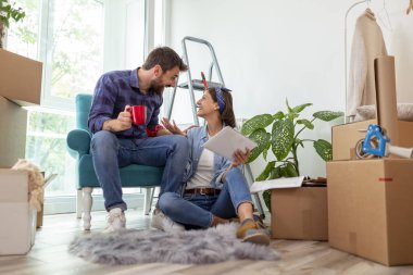 Birbirine aşık bir çift, tablet bilgisayar ve dergiler kullanarak ev dekorasyonu fikirleri ararken kahve içip eğleniyor.