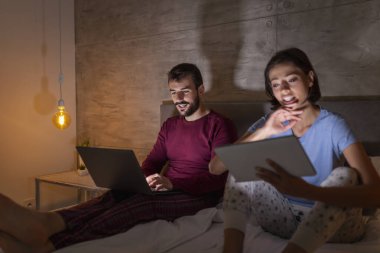 Güzel genç çift geceleri yan yana oturup dizüstü bilgisayar ve tablet bilgisayarlarda TV programları okuyor ve izliyorlar.