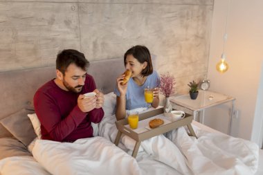 Güzel, genç aşık bir çift pijama giyip yatakta oturuyor, kahve içiyor, kahvaltı yapıyor ve sabah evde dinleniyor.