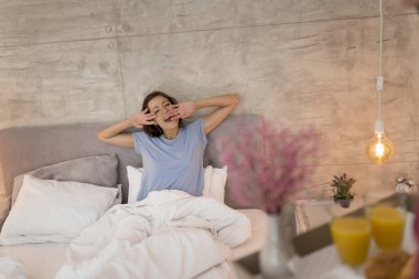 Yatakta pijama giyen güzel bir kız. Erkek arkadaşı kahvaltı ve çiçekleri tepside yatağa getirerek ona sürpriz yapıyor.