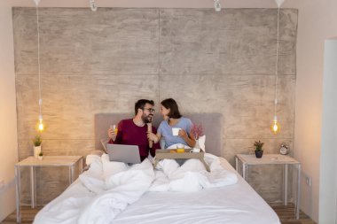 Aşık bir çift yatakta uzanıyor, hafta sonu sabahının tadını çıkarıyor, kahvaltı yapıyor ve internette dizüstü bilgisayarla sörf yapıyor.