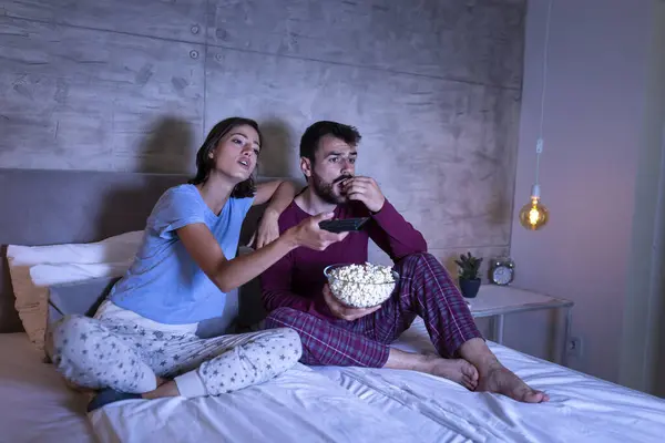 Beautiful young couple sitting in bed, wearing pajamas, eating popcorn and changing channels on TV, searching for a movie or TV show to watch