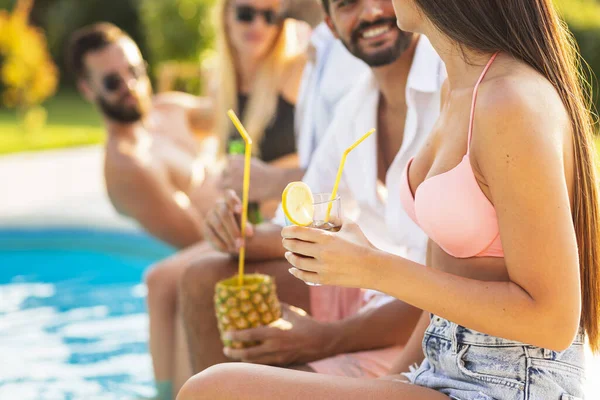 Group of friends at a poolside summer party, sitting at the edge of a swimming pool, drinking cocktails and beer and having fun