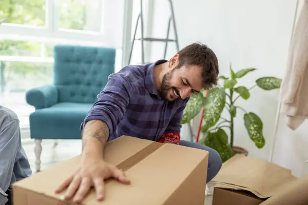 Couple in love packing things into cardboard boxes, getting ready for relocation - man taping boxes using packing machine while woman is wrapping fragile things into paper