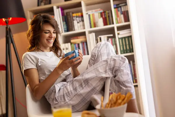 Beautiful young woman wearing pajamas, sitting next to the living room window, holding a cup of coffee and enjoying the moment