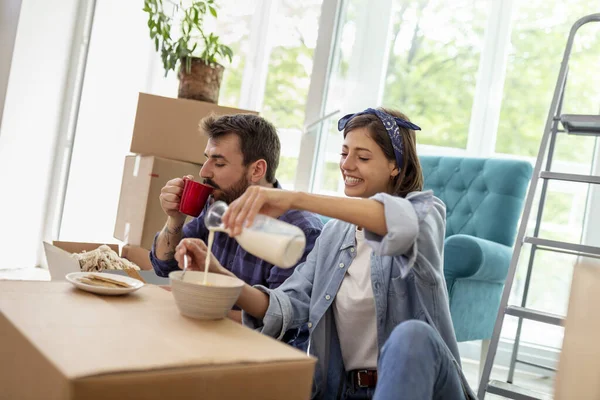 美しい若いカップルは一緒に移動し 新しいアパートの段ボール箱の間に座って 朝食を食べながら楽しんでいます — ストック写真