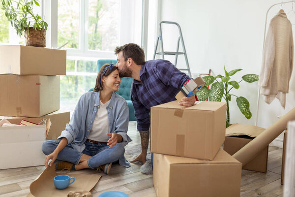 Beautiful young couple in love sitting on the floor of their apartment, kissing and hugging while packing things into cardboard boxes, getting ready for relocation