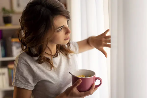 Beautiful young woman wearing pajamas, standing next to the living room window, holding a bowl of breakfast cereals and enjoying the weekend morning