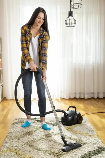 Genç bir kadın ev işleri yapıyor, oturma odasının halısını süpürüyor.