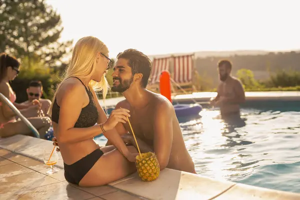 Couple in love at a poolside summer party, sitting at the edge of a swimming pool, sunbathing, drinking cocktails and having fun