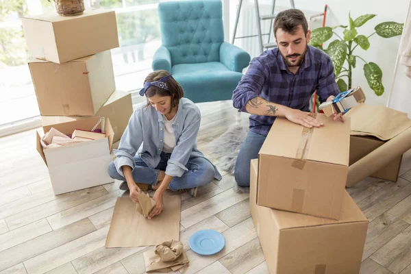 Couple in love packing things into cardboard boxes, getting ready for relocation - man taping boxes using packing machine while woman is wrapping fragile things into paper