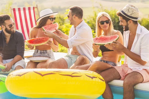 Havuz başında bir yaz partisinde bir grup arkadaş, yüzme havuzunun kenarında oturmuş, soğuk karpuz dilimleri yiyor ve eğleniyorlar.