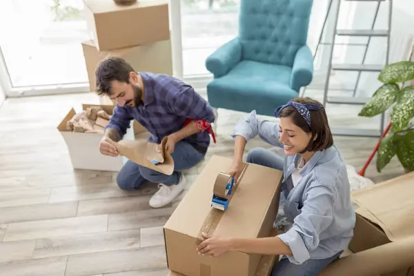 Beautiful young woman using a packing machine, taping cardboard boxes, getting ready for relocation; couple packing things while moving house