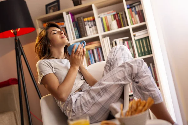 Pijama giyen güzel genç bir kadın, oturma odasının penceresinin yanında oturuyor, elinde bir fincan kahve tutuyor ve anın tadını çıkarıyor.