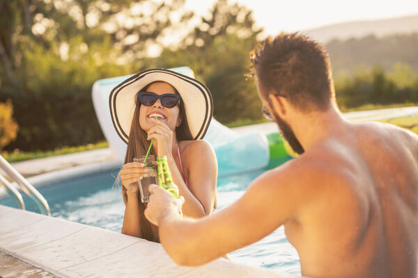 Couple in love at a poolside summer party, standing in water next to the swimming pool edge, drinking beer and having fun