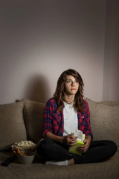 Beautiful young woman sitting in the dark on a living room couch, watching drama movie
