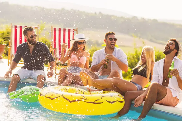 Group of friends at a poolside summer party, sitting at the edge of a swimming pool, drinking beer, splashing water and having fun. Focus on the guy on the right