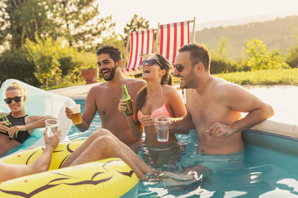 Group of friends at a poolside summer party, having fun in the swimming pool, drinking cocktails and beer and making a toast