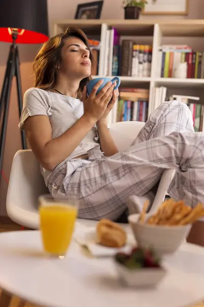 Beautiful young woman wearing pajamas, sitting next to the living room window, holding a cup of coffee and enjoying the moment