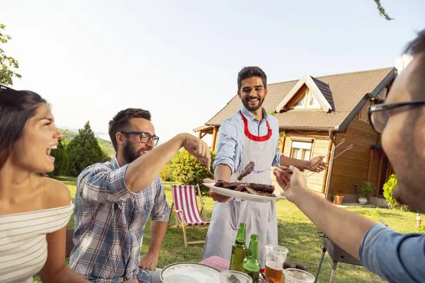 Bir grup arkadaş dışarıda barbekü yapıyor, ızgara et yiyor, bira içiyor ve eğleniyorlar.