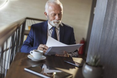 Restoran masasında oturan kıdemli işadamı, belgeleri ve sözleşmeleri analiz ediyor, notlar alıyor ve kahve içiyor.