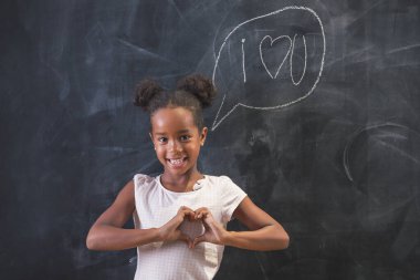 Güzel, küçük melez bir ilkokul öğrencisi sınıfın tahtası önünde duruyor. Elleriyle kalp işaretini gösteriyor ve gülümsüyor.