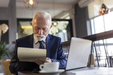 Restoran masasında oturan kıdemli işadamı dizüstü bilgisayarla çalışıyor, anlaşmaları analiz ediyor, imzalıyor ve kahve içiyor.