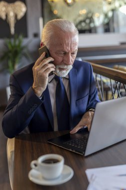 Modern bir ofiste çalışan kıdemli işadamı, dizüstü bilgisayarla uğraşırken telefon görüşmesi yapıyor.