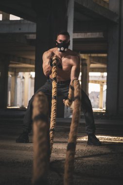 Atletik, kaslı, maske takan, savaş halatları takan, halatlarla dalgalar yaratan, güç toplayan bir adam.