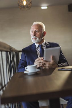 Restoran masasında oturan, kahve içen ve tablet bilgisayarda çalışan kıdemli bir işadamı.