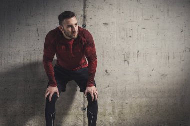 Spor kıyafetleri giyen kaslı atletik bir adam, antrenman molası veriyor, beton bir duvara yaslanıyor, dalgın