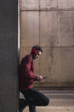 Spor kıyafetleri giyen kaslı estetik bir adam, beton bir duvara yaslanmış, akıllı telefon çalma listesinden bir şarkı seçiyor, egzersiz yaparken müzik dinliyor.