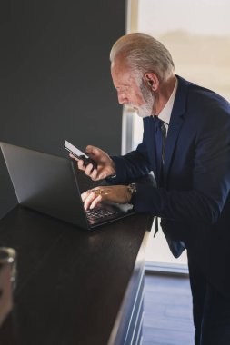 Modern bir ofiste çalışan kıdemli bir işadamı, dizüstü bilgisayar üzerinde çalışırken akıllı bir telefon kullanıyor.