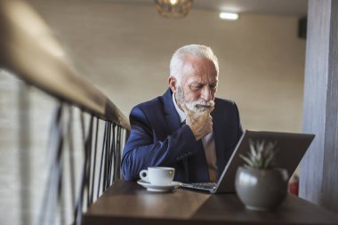 Restoran masasında oturan kıdemli işadamı, bir fincan kahve içiyor ve dizüstü bilgisayardan haber okuyor.