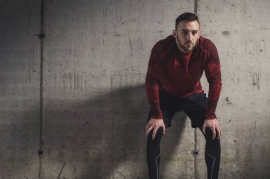 Spor kıyafetleri giyen kaslı atletik bir adam, antrenman molası veriyor, beton bir duvara yaslanıyor, dalgın