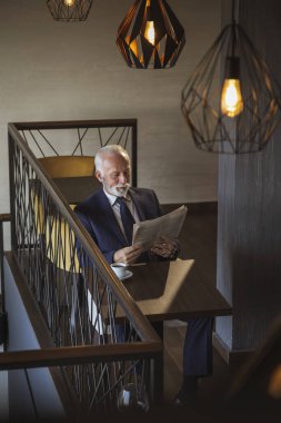 Restoran masasında oturan kıdemli işadamı gazete okuyor ve kahve içiyor.