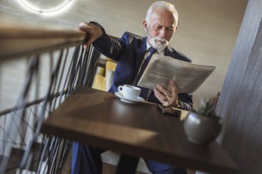 Üst düzey işadamı modern bir ofis binasında oturuyor, kahve içiyor ve gazete okuyor.