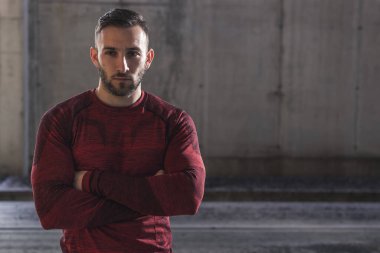 Spor kıyafetleri giyen kaslı atletik bir adam spor yapıyor, antreman molası veriyor, kolları çaprazlanmış beton bir duvarın önünde kameraya bakıyor.