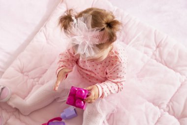 Pembe etek giyen, yorganın üzerinde oturan ve oyuncak bloklarla oynayan bir kızın yüksek açılı görüntüsü.