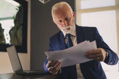 Modern bir ofiste kıdemli bir işadamı dizüstü bilgisayar üzerinde çalışıyor ve sözleşmeleri ve belgeleri analiz ediyor, ciddi ve düşünceli.
